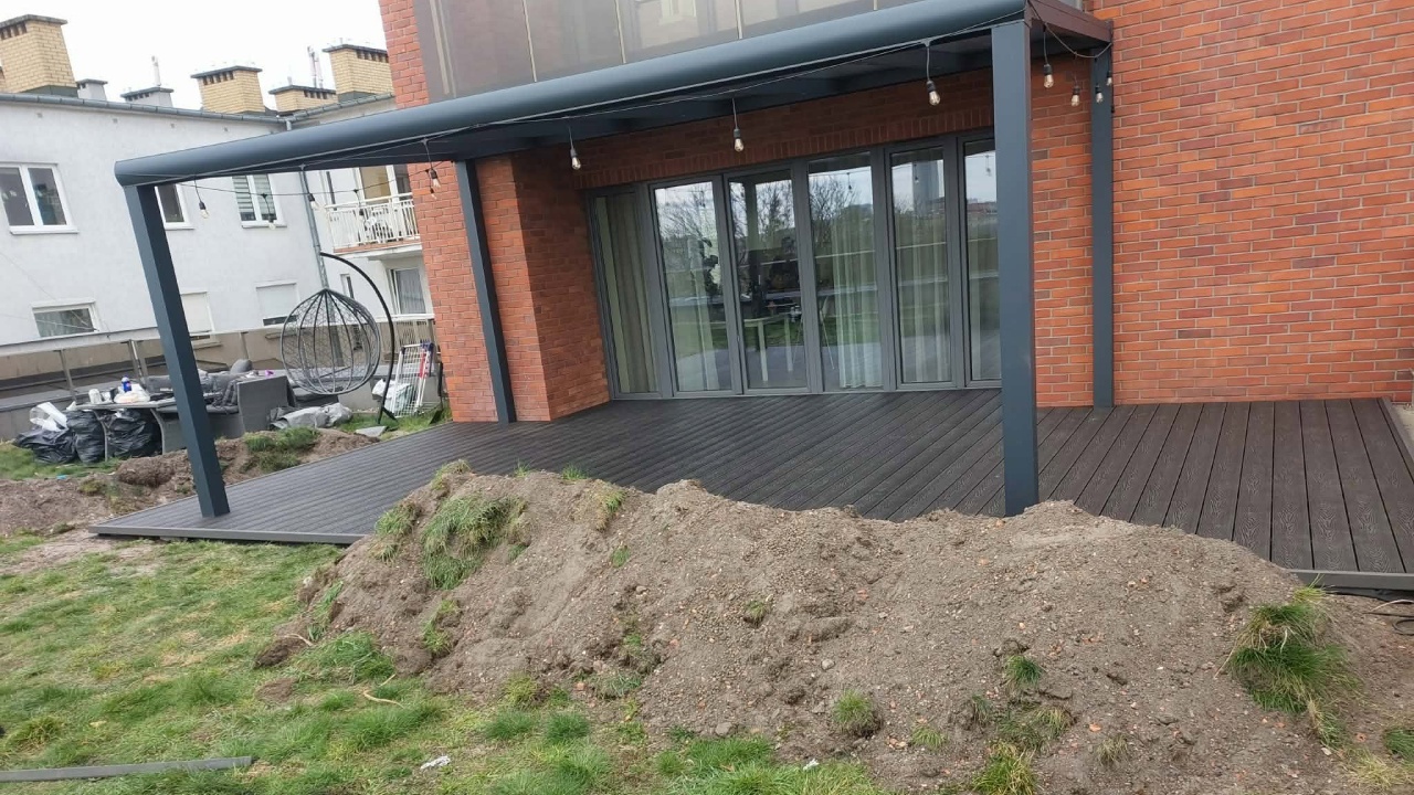 Taras z grafitowych desek kompozytowych pod ciemnym zadaszeniem przy budynku z czerwonej cegły.