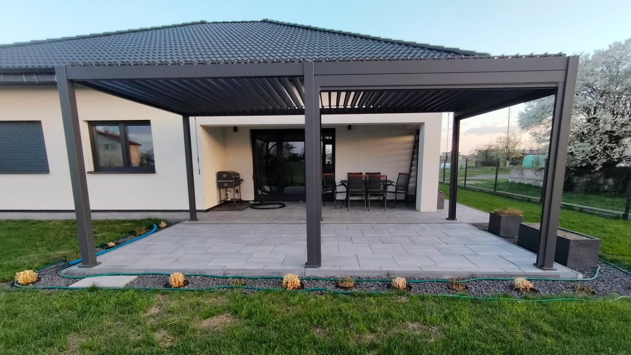 Ciemnoszara pergola lamelowa zadaszająca szary taras przy domu jednorodzinnym z meblami ogrodowymi.